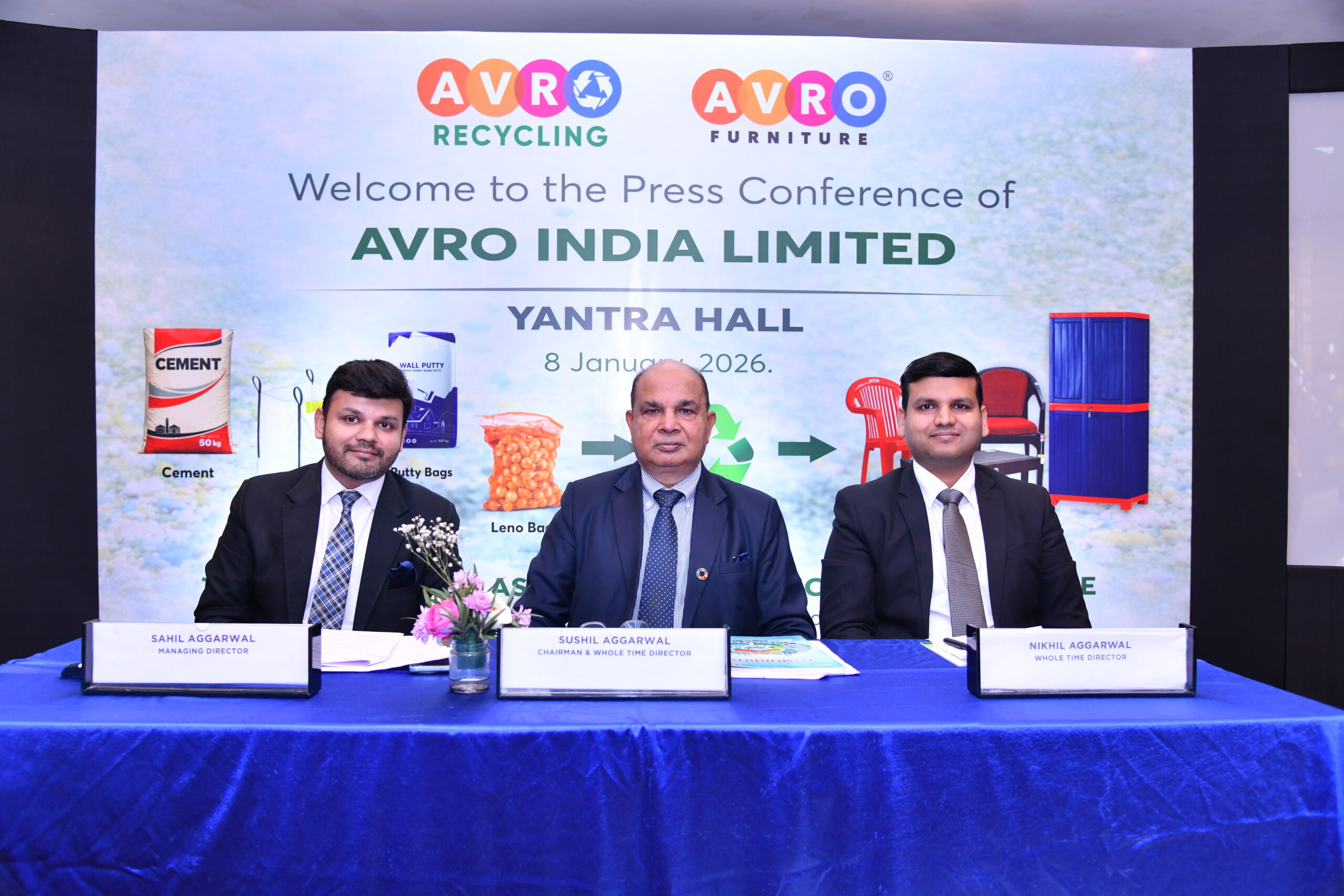 Three men in formal business suits sit behind a blue-covered table at a press conference for AVRO India Limited, held at Yantra Hall on 8 January 2026. The backdrop displays the AVRO Recycling and AVRO Furniture logos along with the event title and graphics illustrating plastic waste being transformed into furniture products. Nameplates identify the speakers as Sahil Aggarwal (Managing Director), Sushil Aggarwal (Chairman & Whole Time Director), and Nikhil Aggarwal (Whole Time Director). A small flower arrangement sits on the table in front of them.